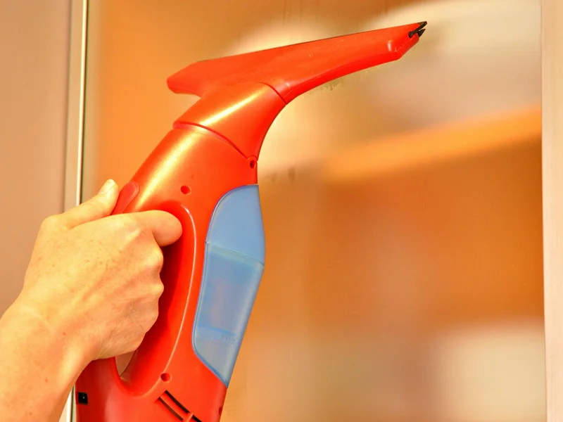 Main tenant un appareil rouge sur une surface vitrée — nettoyeur vapeur surfaces