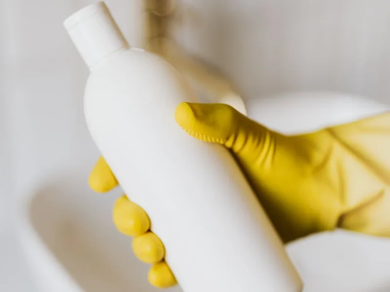 Main gantée jaune tenant une bouteille de produit nettoyant dans un lavabo blanc, pour illustrer l'utilisation du savon noir sur les sanitaires