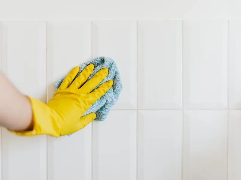 Main gantée jaune avec microfibre bleue nettoyant du carrelage blanc de salle de bain, illustrant le protocole savon noir sur les sanitaires