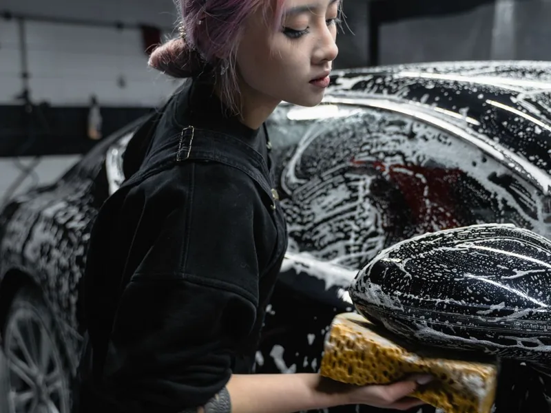 Personne avec une éponge jaune lavant la carrosserie d'une voiture noire couverte de mousse, en atelier de lavage