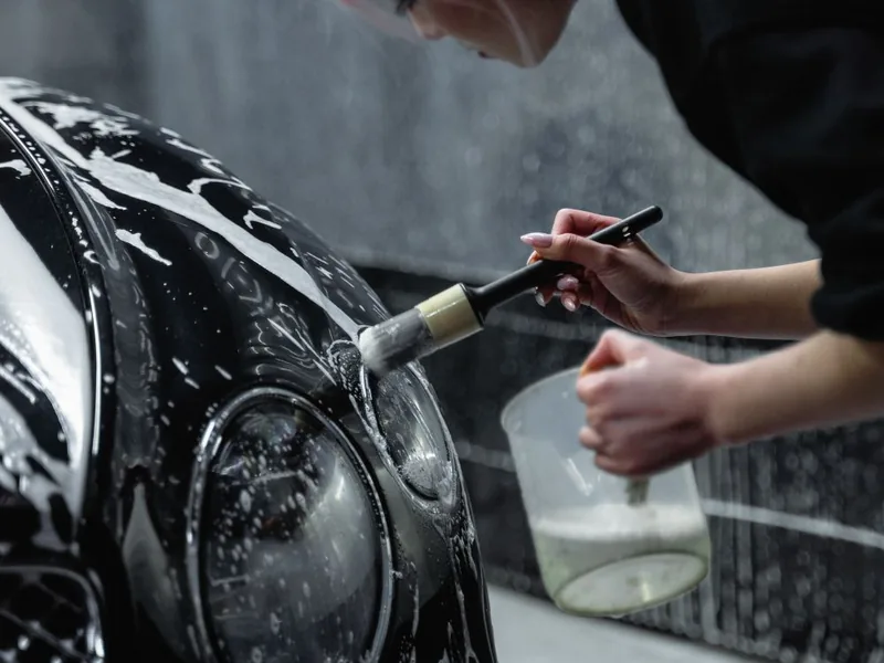 Gros plan d'une main appliquant un pinceau avec du produit savonneux sur la carrosserie noire d'une voiture, illustrant le nettoyage des zones de jonction